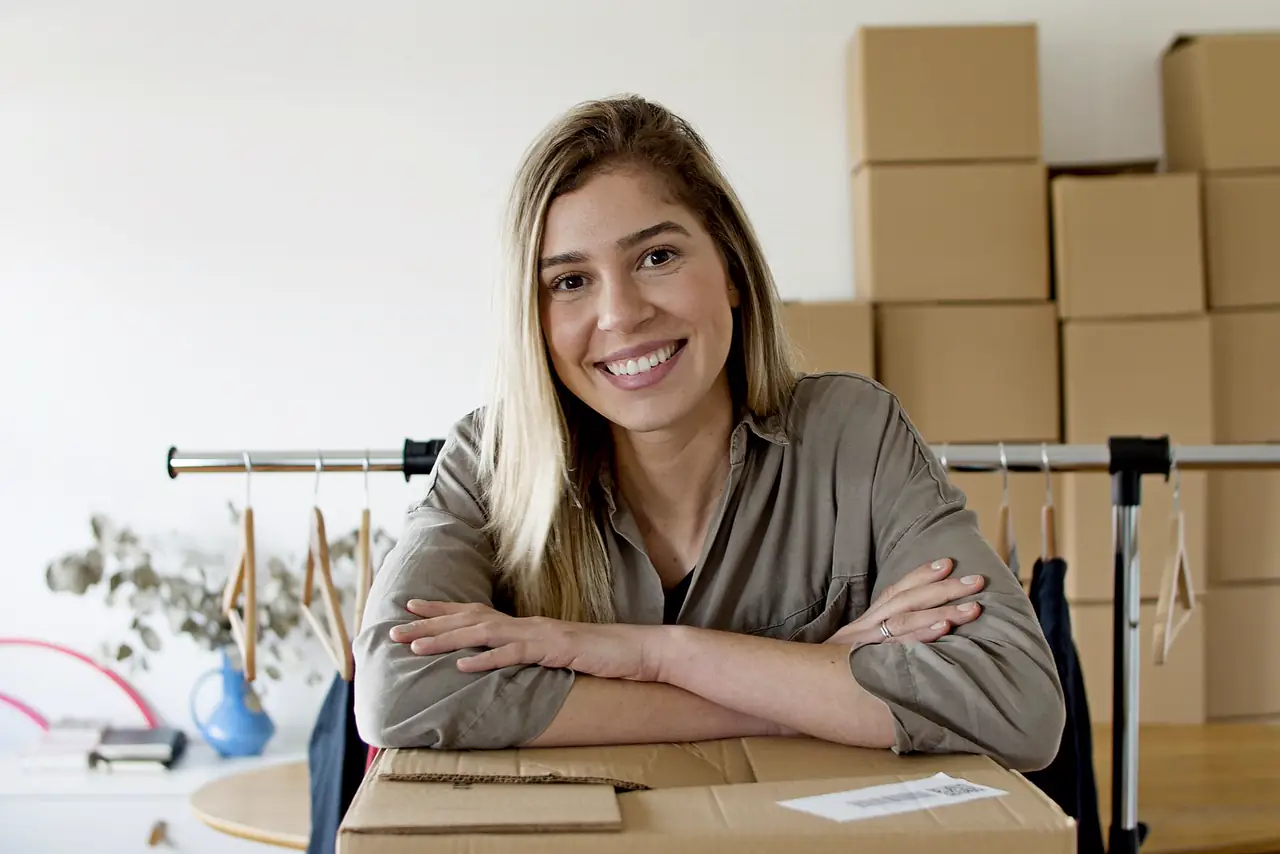 Happy online shop owner sits at her packing desk beside parcels and a clothing rail