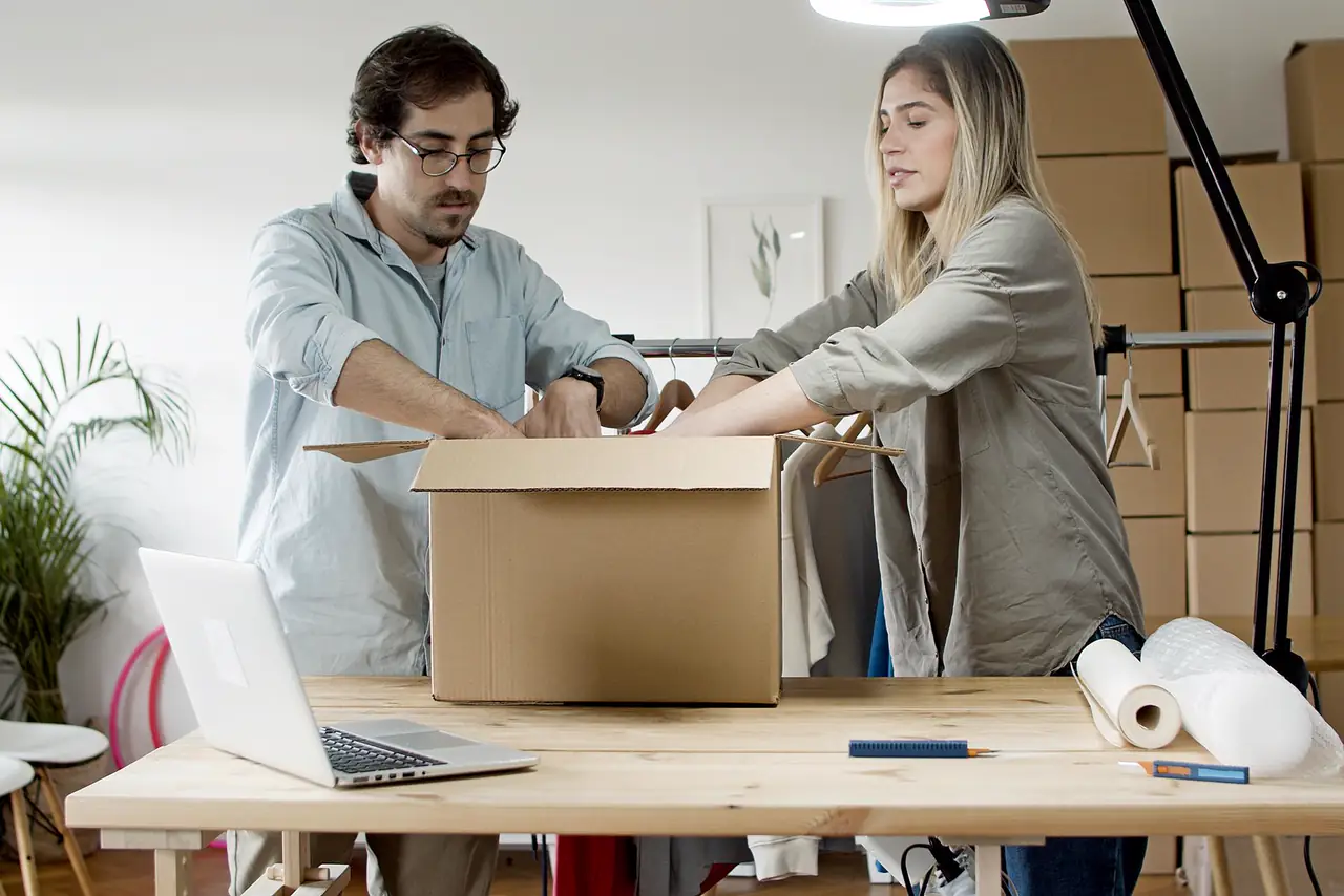 Two sellers pack customer orders into a box beside a laptop and parcels