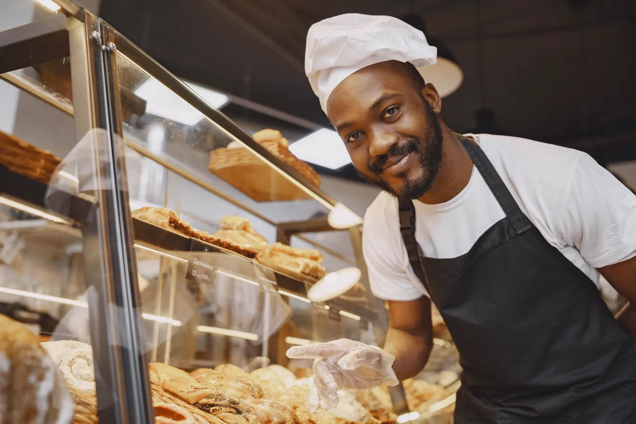 Smiling baker in a white cap and apron arranges fresh pastries behind a glass bakery counter