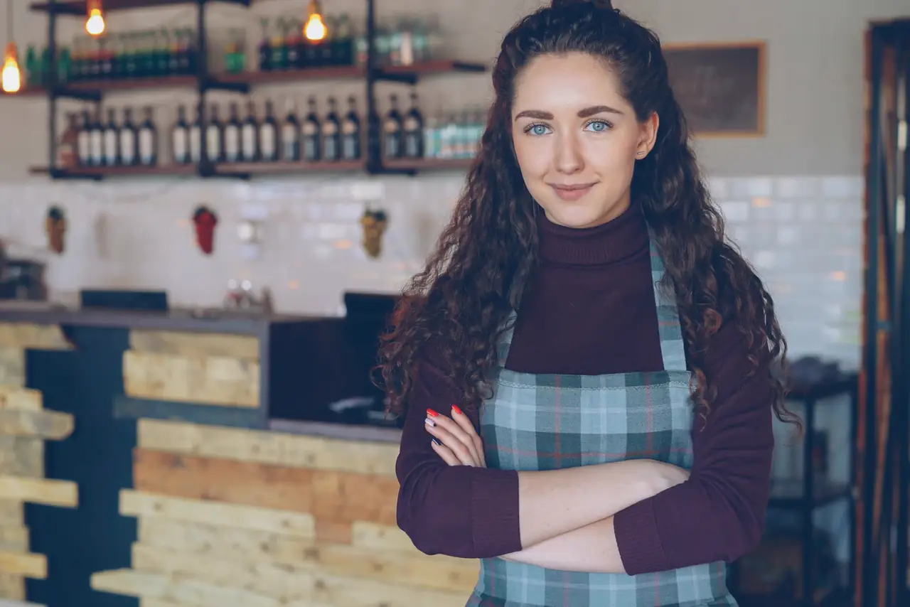 Confident cafe owner in an apron stands with arms crossed inside a rustic coffee shop