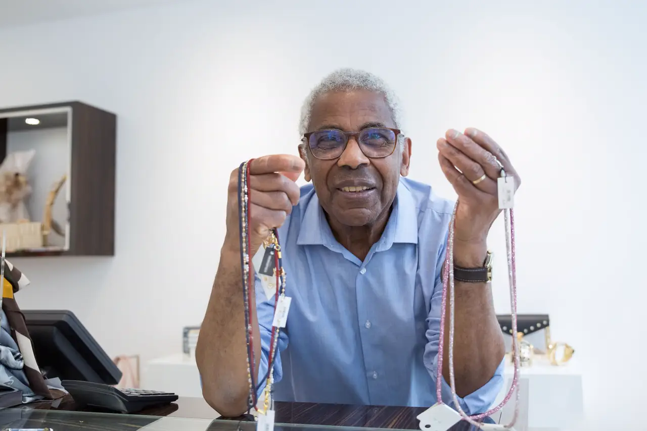 Smiling senior jeweller holds up necklaces in his bright shop
