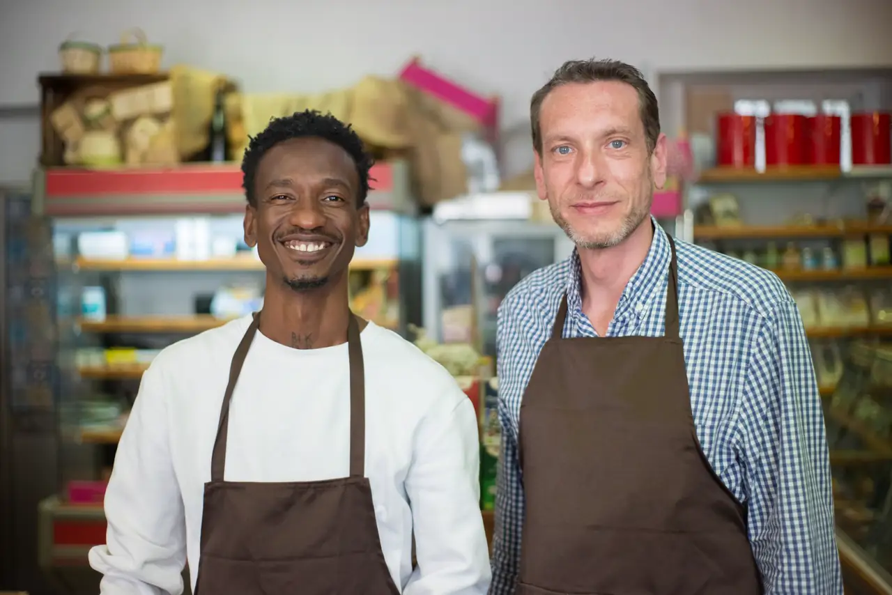 Two happy grocery store owners in aprons stand together in their shop