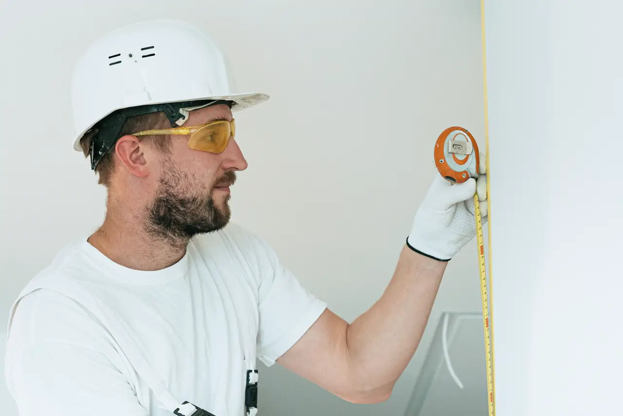 Construction worker in a hard hat and goggles measures a wall with a tape measure